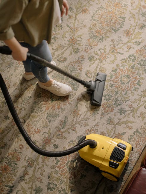 A person wearing beige shoes, blue jeans, and a beige jacket is using a vacuum cleaner to clean a patterned area rug in a room with soft, warm lighting. The vacuum cleaner is yellow with a black hose, and the person is holding a wand attachment over the rug surface, which features a floral design in muted tones of green, orange, and cream. The space appears tidy, with the rug covering a wooden floor, and part of a piece of furniture is visible in the corner. The scene suggests domestic deep cleaning carried out by Chelsea Carpet Cleaning to maintain hygiene and surface cleanliness.