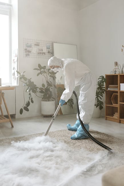 A person wearing beige shoes, blue jeans, and a beige jacket is using a vacuum cleaner to clean a patterned area rug in a room with soft, warm lighting. The vacuum cleaner is yellow with a black hose, and the person is holding a wand attachment over the rug surface, which features a floral design in muted tones of green, orange, and cream. The space appears tidy, with the rug covering a wooden floor, and part of a piece of furniture is visible in the corner. The scene suggests domestic deep cleaning carried out by Chelsea Carpet Cleaning to maintain hygiene and surface cleanliness.