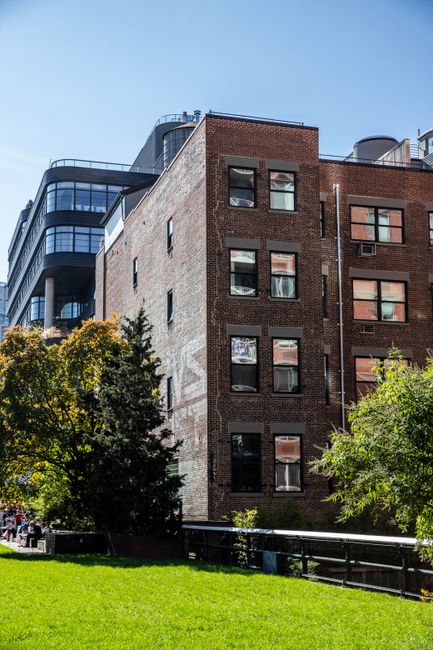 Exterior view of a modern multi-story residential building on the Cadogan Estate in Chelsea, featuring a combination of brick and glass facades. The brick section has a traditional appearance with several rectangular windows, some reflecting the surrounding sky and trees. Adjacent to it, the glass section exhibits curved, dark-tinted windows and balconies, emphasizing contemporary architectural design. Surrounding the building, there are lush, green trees and a well-maintained grassy area, indicating a landscaped outdoor space. The scene is brightly lit under clear daylight, highlighting the clean and well-kept exterior, consistent with high standards of property maintenance and hygiene promoted by Chelsea Carpet Cleaning for end-of-tenancy cleaning in Chelsea.