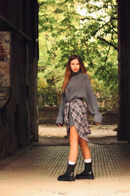 A young woman with long brown hair stands at the entrance of a rustic wooden shed or barn, with an open door revealing lush green trees and foliage in the background. She is dressed in a gray oversized knit sweater, a plaid shirt tied around her waist, black work boots, and white socks. The ground beneath her is covered in textured wooden planks, and the scene is illuminated by natural daylight, creating a warm and inviting atmosphere. The setting appears to be outdoors, surrounded by nature, with no visible cleaning tools or surfaces present. This image does not directly relate to surface cleaning or deep cleaning but could be used to illustrate outdoor or rustic cleaning themes as seen in the context of Chelsea Carpet Cleaning's services.