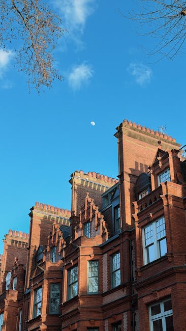Exterior view of a modern multi-story residential building on the Cadogan Estate in Chelsea, featuring a combination of brick and glass facades. The brick section has a traditional appearance with several rectangular windows, some reflecting the surrounding sky and trees. Adjacent to it, the glass section exhibits curved, dark-tinted windows and balconies, emphasizing contemporary architectural design. Surrounding the building, there are lush, green trees and a well-maintained grassy area, indicating a landscaped outdoor space. The scene is brightly lit under clear daylight, highlighting the clean and well-kept exterior, consistent with high standards of property maintenance and hygiene promoted by Chelsea Carpet Cleaning for end-of-tenancy cleaning in Chelsea.
