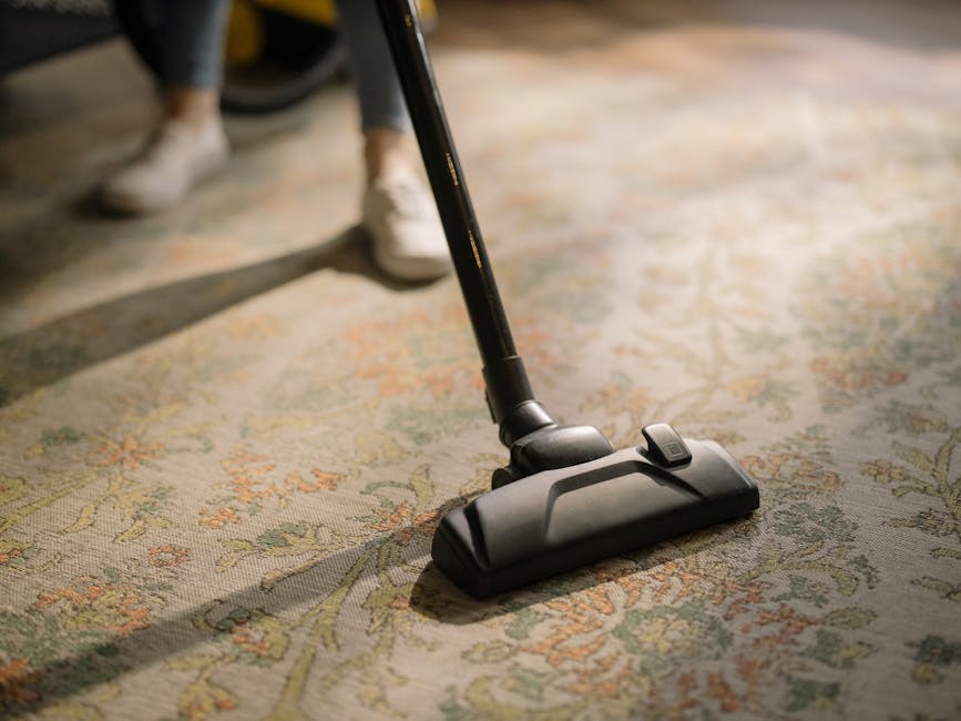 Close-up of a person's white sneakers standing on an ornate floral patterned area rug in a well-lit room. A black vacuum cleaner with a slim nozzle attachment is being used to deep clean the rug's surface, which appears clean and free of dust. The person's legs are visible from the knees down, with the rest of the room out of focus. The scene emphasizes surface cleaning and maintenance, showcasing tools used in professional domestic cleaning services provided by Chelsea Carpet Cleaning, specifically in the Chelsea SW3 area as highlighted on the webpage.