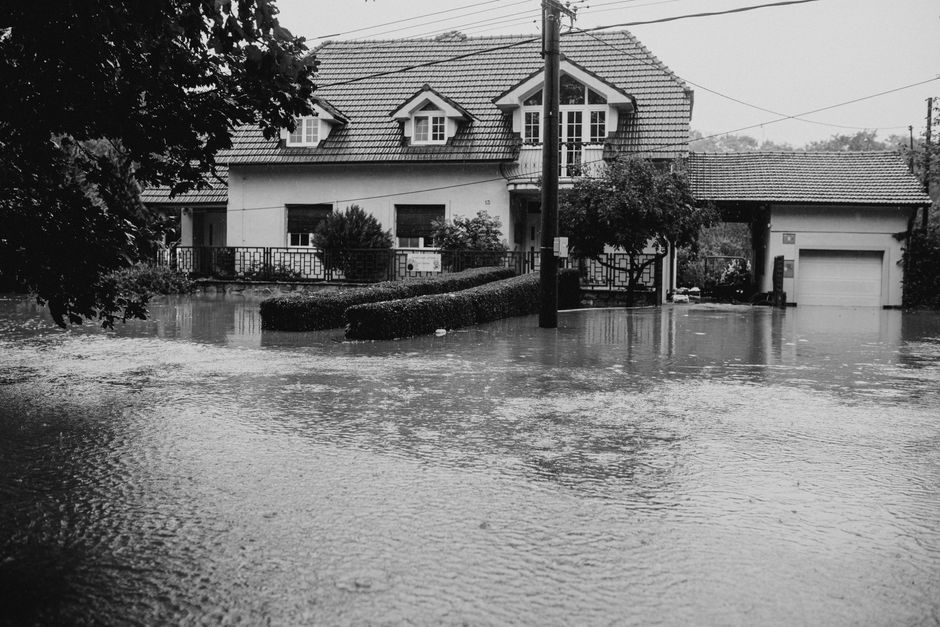 A two-story residential house with a tiled gabled roof and white exterior walls, surrounded by a garden with bushes and a small tree. The house's front yard is heavily flooded, with water covering the ground up to the porch area and extending across the driveway, which includes a closed garage door on the right. A wooden fence encloses the garden, and an electric pole stands in front of the house. The scene is captured in black and white, highlighting the extent of water damage caused by flooding, which is relevant to water damage emergency services provided by Chelsea Carpet Cleaning in Chelsea. Although the image does not display any cleaning tools, the context relates to water damage cleanup and deep cleaning after flooding incidents.