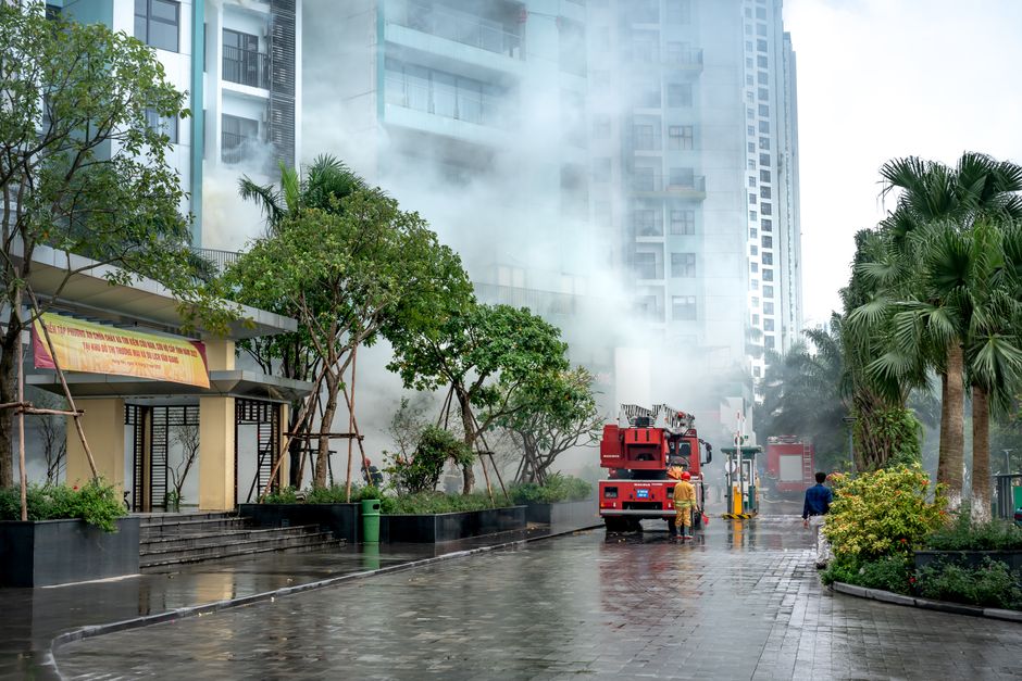 An outdoor scene near a modern high-rise building featuring a fire truck with a mounted ladder and firefighters spraying water. The wet pavement reflects the surroundings, and smoke is visibly emanating from the building, suggesting an emergency response to water damage or a fire. Green trees and landscaped bushes line the sidewalk, with some scaffolding or supports visible on the left side. The area appears to be a commercial or residential complex, and the lighting indicates daytime with overcast skies. The image illustrates the active use of cleaning and safety procedures, consistent with emergency water damage management, as handled by Chelsea Carpet Cleaning, which specializes in deep cleaning and sanitisation services.