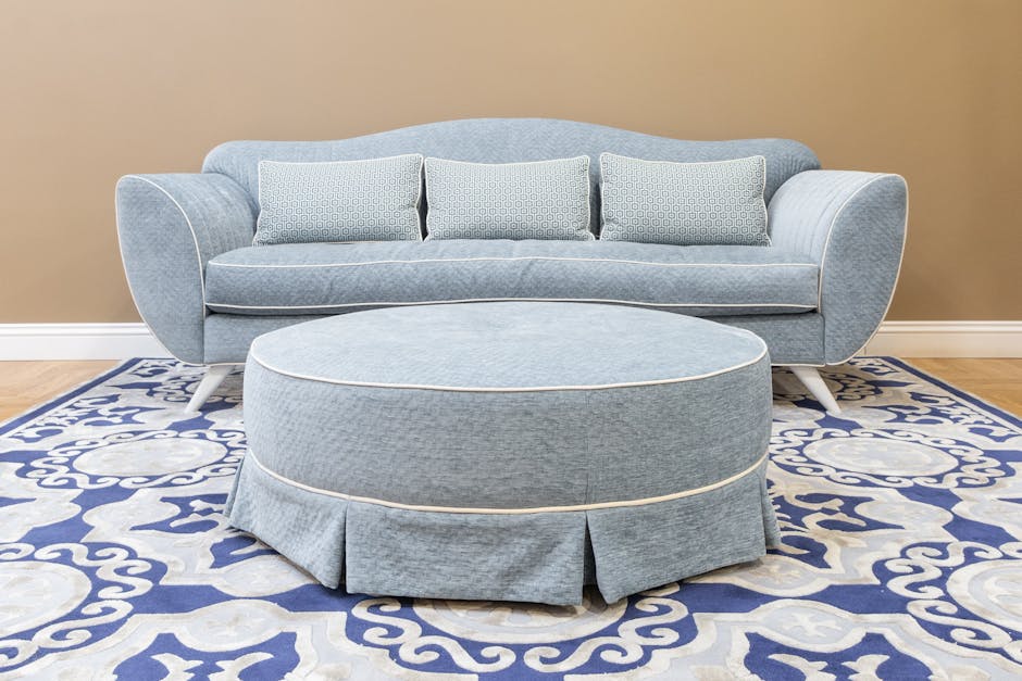 A light blue upholstered sofa with curved armrests and three matching cushions, positioned against a beige wall in a well-lit room. In front of the sofa, a round, matching light blue ottoman with a pleated skirt is placed on a patterned blue and white rug. The room features a wooden floor, and the overall appearance shows a clean and tidy seating area, indicative of professional domestic cleaning or surface cleaning by Chelsea Carpet Cleaning, with a focus on maintaining upholstery hygiene and appearance.