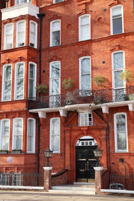 Exterior view of a red brick residential building with multiple bay windows and black wrought iron balconies. The facade features white window frames and decorative brickwork details. Two small trees are planted in front of the building, along with a black wrought iron fence and traditional street lamps at the entrance. The lighting suggests daytime with overcast skies, emphasizing the building's classic appearance and well-maintained exterior. This image highlights the architecture and urban setting typical of Sloane Square, with Chelsea Carpet Cleaning specializing in surface cleaning and deep cleaning services for homes in the Chelsea area.